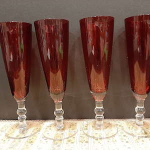 Könnte beinhalten: Set aus vier roten Glas-Champagnerflöten mit klaren Stielen und Füßen. Die Flöten sind in einer Reihe auf einer gemusterten Oberfläche angeordnet.