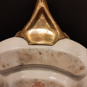 May include: A white ceramic serving dish with a gold-colored handle and a red and gold logo with a crown and the text "1895".