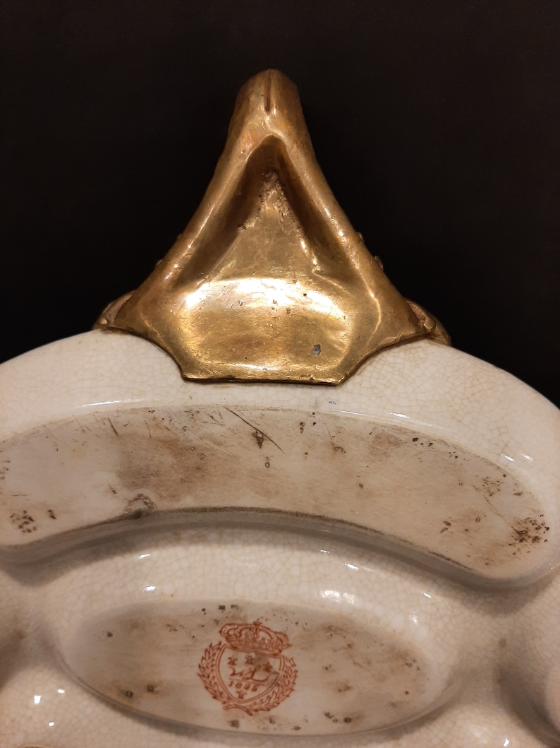 May include: A white ceramic serving dish with a gold-colored handle and a red and gold logo with a crown and the text "1895".