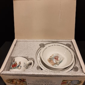 Może przedstawiać: Zestaw naczyń dla dzieci Peter Rabbit w pudełku. Zestaw zawiera biały kubek i dwie białe miseczki z ilustracjami Petera Rabbit i tekstem. Pudełko jest otwarte, ukazując zestaw umieszczony w szarej wkładce z pianki.