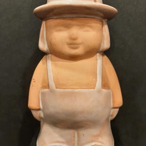 May include: A ceramic figurine of a person wearing a hat and overalls. The figurine is made of terracotta and has a light brown glaze. The person has a simple, smiling face.