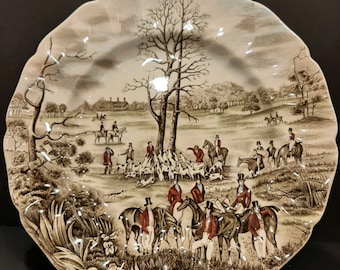 May include: A brown and white ceramic plate with a hunting scene. The plate features a group of people on horseback riding through a forest with hounds. The plate has a scalloped edge.