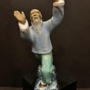 Op de afbeelding: Een keramiek beeldje van een oudere man met een lange witte baard, gekleed in een blauwe en groene robe, in een tai chi pose. Het beeldje staat op een rechthoekige basis met een blauwe en witte glazuur.