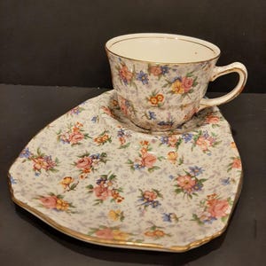 Könnte beinhalten: Ein weißes Tee-Set mit Untertasse mit einem Blumenmuster in Rosa-, Blau- und Gelbtönen. Das Set hat einen goldenen Rand.
