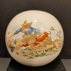 May include: A round, off-white decorative sphere featuring a colorful illustration of two rabbits running across a field. One rabbit wears a blue jacket and yellow hat, the other an orange vest. A small mouse sweeps the ground.