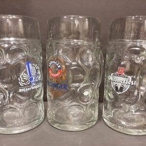 May include: Three clear glass beer steins with textured, bubble-like patterns. Each stein has a different logo: "Steam Whistle", "Erdinger Weissbrau", and "Richard's Oktoberfest". The steins are designed with a handle and a thick base.
