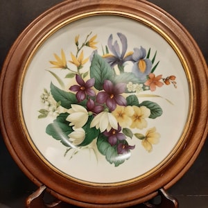Könnte beinhalten: Ein dekorativer Teller mit floralem Design, mit lila, gelben und weißen Blumen, in einem Holzrahmen. Der Teller wird auf einem Holzständer präsentiert und zeigt die komplizierten Details der botanischen Kunstwerke.