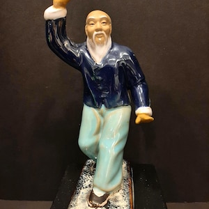 Op de afbeelding: Een keramiek beeldje van een man met een lange witte baard, die een blauw jasje en een lichtblauwe broek draagt. Hij staat op een kleine voet en heeft zijn rechterarm omhoog.