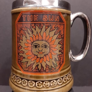 Pode incluir: Uma caneca de cerâmica marrom com tampa e alça prateadas. A caneca tem um gráfico de um sol com rosto e as palavras "THE SUN" na frente.