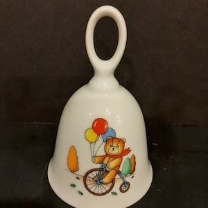 Könnte beinhalten: Weiße Keramikglocke mit einem Henkel in Form einer Schlaufe. Die Glocke zeigt eine bunte Illustration eines Teddybären, der ein Fahrrad mit Ballons fährt.