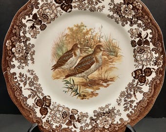 May include: A decorative plate with a brown floral border and a central image of two birds in a natural setting. The plate has a scalloped edge and is supported by a black stand. The color palette includes shades of brown, beige, and white.