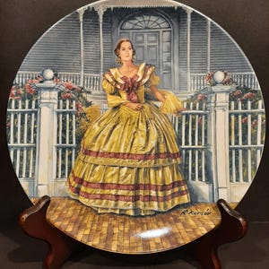 May include: A decorative plate featuring a woman in a yellow dress with red accents, standing in front of a white fence and house. The plate is displayed on a wooden stand, showcasing a detailed painting of a historical scene.