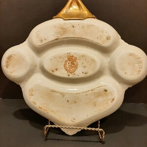 May include: White ceramic dish with four compartments and a gold-colored handle. The dish has a brown stamp with a crown and the text "Royal Copenhagen" in the center.