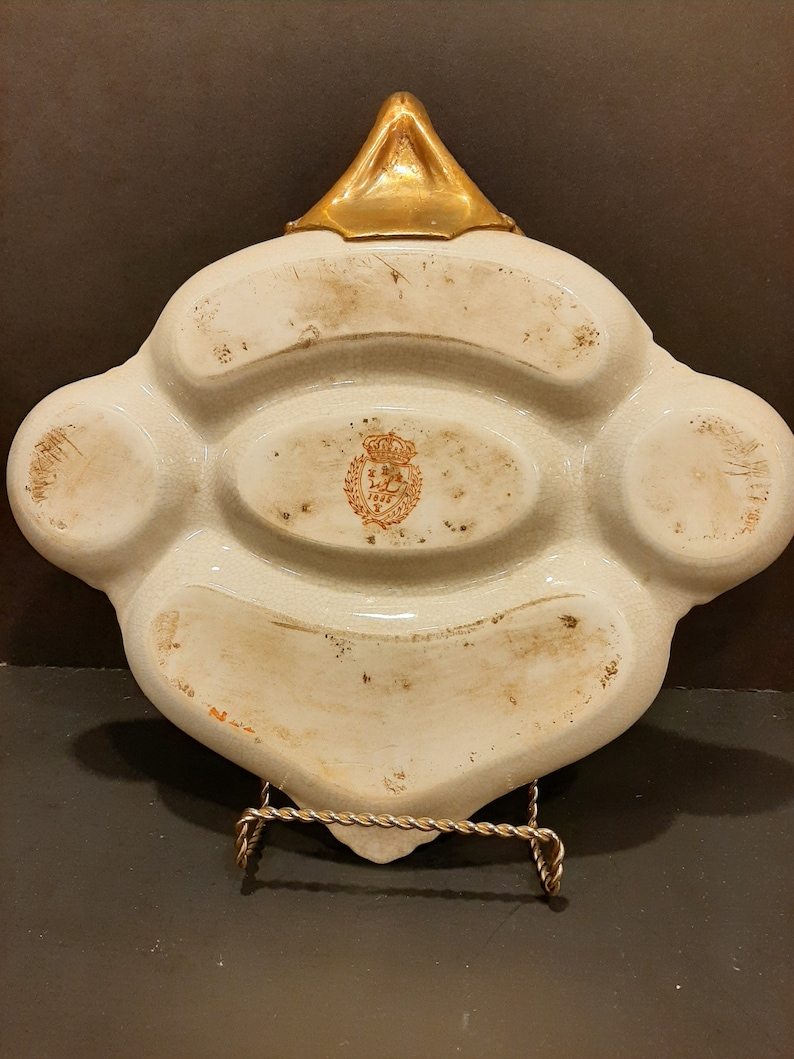 May include: White ceramic dish with four compartments and a gold-colored handle. The dish has a brown stamp with a crown and the text "Royal Copenhagen" in the center.