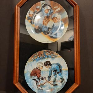 May include: A framed set of decorative plates depicting hockey scenes. The top plate shows young hockey players in blue uniforms. The bottom plate shows two players in action, one in a maroon jersey and the other in blue. The frame is a dark wood octagon shape.
