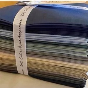 May include: A stack of fabric squares in various shades of blue, green, beige, and brown. The fabric is neatly folded and tied with a white ribbon that says "Cut out for happiness".