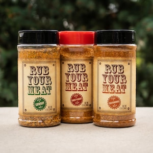 May include: Three clear plastic spice bottles with black and red lids. Each bottle has a label that reads "RUB YOUR MEAT" in a vintage font. The labels also include flavor names: "Cowboy", "Sweet 'N Sassy", and "Old Fashioned".