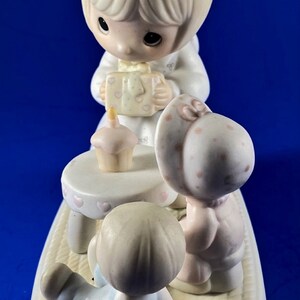 以下が含まれることがあります： 誕生日を祝う3人の子供たちの磁器製フィギュア。女の子がプレゼントを持っており、男の子が小さなテーブルに座り、女の子がカップケーキを持っています。フィギュアは白で、ピンクとブルーのアクセントが付けられています。
