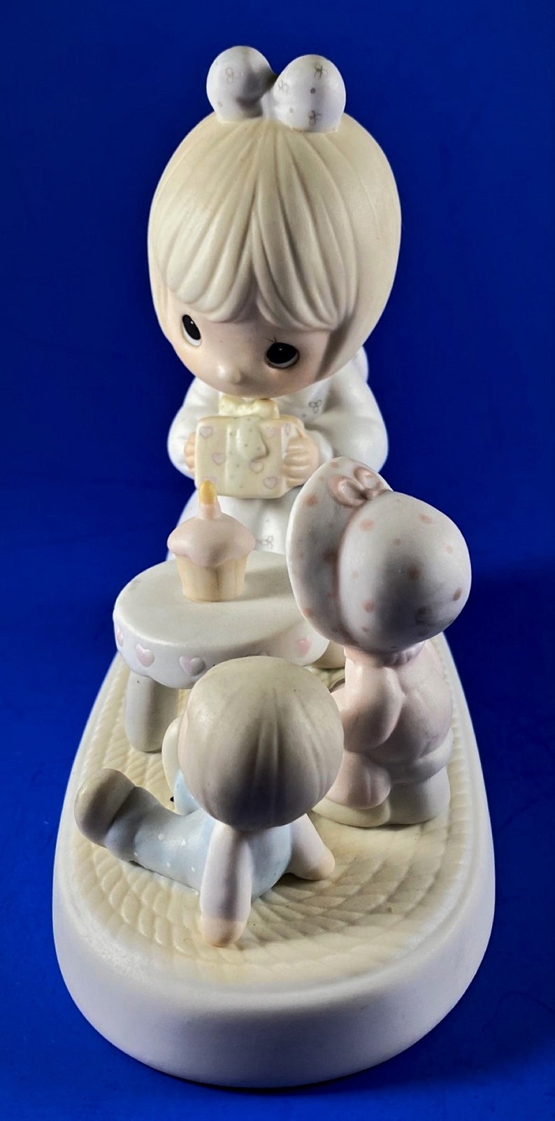 以下が含まれることがあります： 誕生日を祝う3人の子供たちの磁器製フィギュア。女の子がプレゼントを持っており、男の子が小さなテーブルに座り、女の子がカップケーキを持っています。フィギュアは白で、ピンクとブルーのアクセントが付けられています。