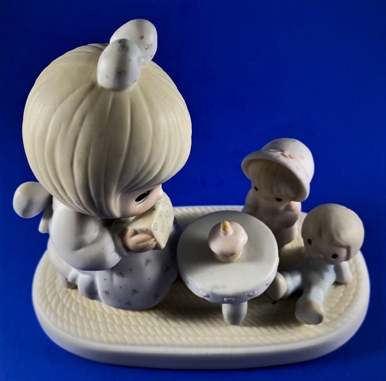 以下が含まれることがあります： 3体のフィギュアがセットになった磁器製の置物です。金髪の女性がプレゼントを持っており、ピンクの帽子をかぶった女の子と青い服を着た男の子がいます。フィギュアは、カップケーキが置かれた小さなテーブルの周りに座っています。置物は、織り模様のついた白い台座の上に置かれています。