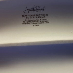 以下が含まれることがあります： 「May your birthday be a blessing」というテキストと、「&copy; 1983 Jonathan & David All rights reserved worldwide Licensee Enesco Imports Corp. E-2826」という著作権情報が書かれた白いセラミック製の人形。