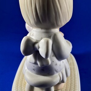 以下が含まれることがあります： 白い花柄のドレスと白いリボンをつけた金髪の女の子の磁器製フィギュア。彼女は、籐製のベースが付いた白い腰掛けに座っています。フィギュアは白いベースの上に置かれています。
