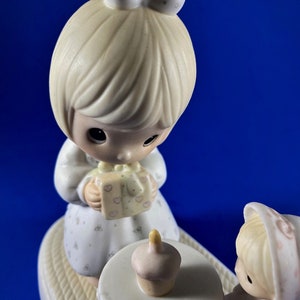 以下が含まれることがあります： 誕生日を祝う2人の女の子の磁器製フィギュア。左側の女の子はピンクのリボンが付いたギフトボックスを持っています。右側の女の子は、カップケーキが置かれた小さなテーブルの隣に座っています。フィギュアはパステルカラーで描かれており、繊細で幻想的なスタイルです。