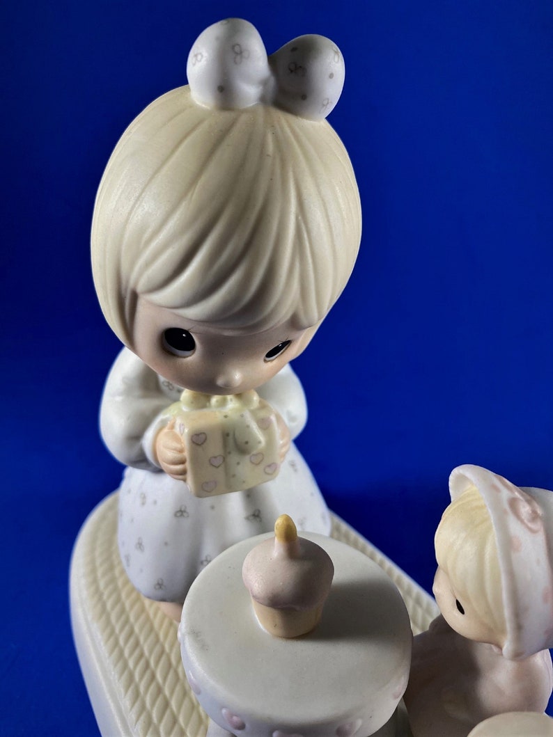以下が含まれることがあります： 誕生日を祝う2人の女の子の磁器製フィギュア。左側の女の子はピンクのリボンが付いたギフトボックスを持っています。右側の女の子は、カップケーキが置かれた小さなテーブルの隣に座っています。フィギュアはパステルカラーで描かれており、繊細で幻想的なスタイルです。