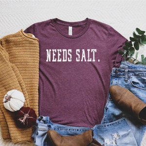 May include: A maroon t-shirt with the text "NEEDS SALT." printed in white. The shirt is laid out on a white surface with a brown sweater, a pair of blue jeans, and brown ankle boots.