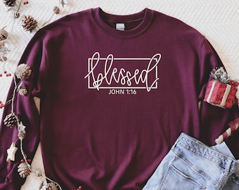 May include: Burgundy sweatshirt with white text that reads "blessed" in a script font with a box around it and "JOHN 1:16" underneath. The sweatshirt is folded and laid on a white surface with blue jeans and a small red gift box.