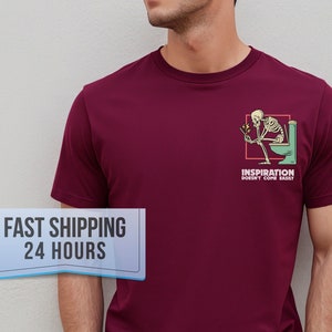 May include: Burgundy t-shirt with a humorous graphic of a skeleton on a toilet, holding a phone. The text "INSPIRATION DOESN'T COME EASILY" is below the graphic. The shirt is a solid color and has short sleeves.