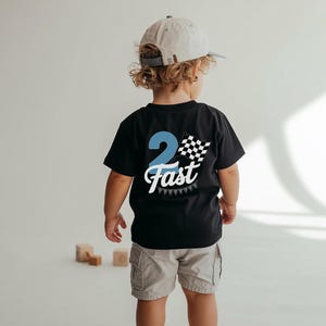 A young child wearing a black t-shirt with a checkered flag design and the number "2 Fast" on the back, standing in front of a white background with wooden blocks.