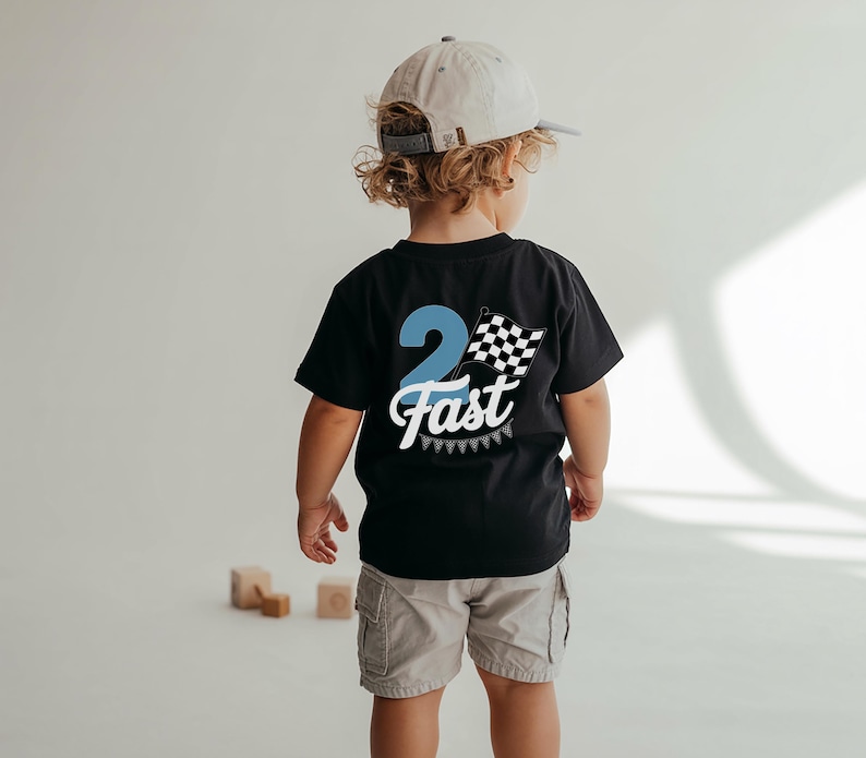 A young child wearing a black t-shirt with a checkered flag design and the number "2 Fast" on the back, standing in front of a white background with wooden blocks.