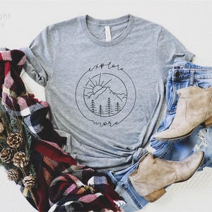 May include: A heather gray t-shirt featuring a black line art graphic of mountains, pine trees, and a sun inside a circle with the text explore more. The shirt is paired with blue denim jeans, tan ankle boots, and a red, black, and white plaid scarf.