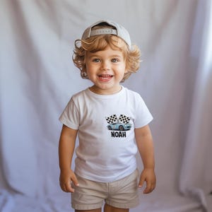 A smiling young child with curly blonde hair wearing a white t-shirt with a graphic of a car and the word "Noah" printed on it.