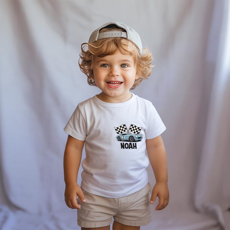 A smiling young child with curly blonde hair wearing a white t-shirt with a graphic of a car and the word "Noah" printed on it.