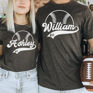 May include: Two grey t-shirts with a white baseball graphic and the names "Ashley #7" and "William #11" printed on them.