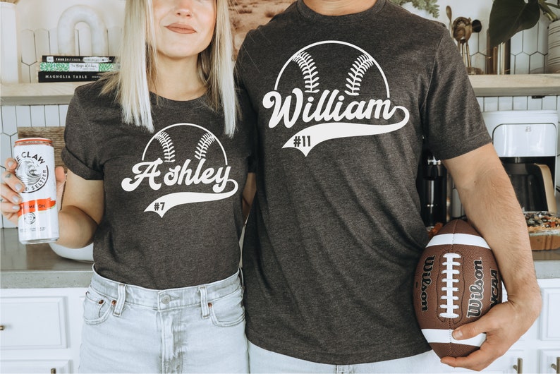 May include: Two grey t-shirts with a white baseball graphic and the names "Ashley #7" and "William #11" printed on them.