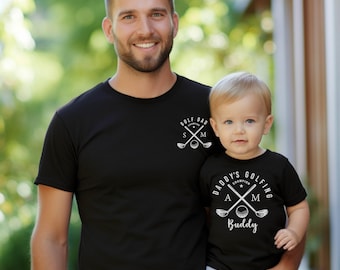 May include: Two black t-shirts featuring white graphic prints. The adult shirt displays crossed golf clubs, a golf ball, and the text GOLF DAD CHAMPION S M. The smaller shirt features crossed golf clubs, a golf ball, and DADDY'S GOLFING BUDDY.