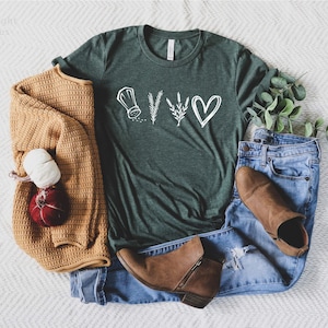May include: A dark green short-sleeve t-shirt featuring a white graphic of a salt shaker, a sprig of rosemary, a sprig of lavender, and a heart. The shirt is paired with a mustard yellow knit cardigan, blue denim jeans, and brown ankle boots.