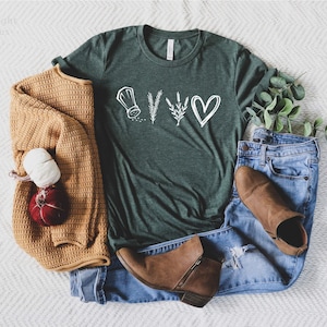 May include: A dark green short-sleeve t-shirt featuring a white graphic of a salt shaker, a sprig of rosemary, a sprig of lavender, and a heart. The shirt is paired with blue denim jeans, a mustard yellow knit cardigan, and brown ankle boots.