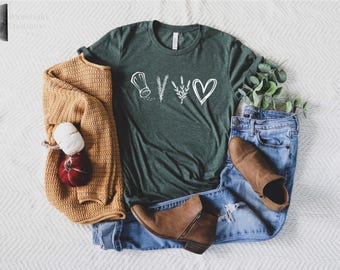 Puede incluir: Una camiseta de manga corta de color verde oscuro con un gráfico blanco de un salero, una rama de romero, una rama de lavanda y un corazón. La camiseta se combina con vaqueros azules, un cárdigan de punto amarillo mostaza y botines marrones.