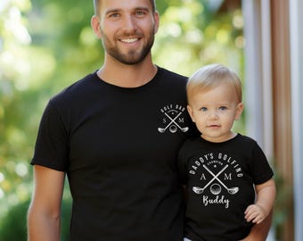 May include: Two black short-sleeved t-shirts with white graphic prints. The adult shirt features crossed golf clubs, a golf ball, and the text GOLF DAD CHAMPION S M. The child shirt features crossed golf clubs, a golf ball, and DADDY'S GOLFING BUDDY.