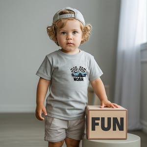 A young child with curly hair wearing a gray t-shirt with a graphic of a car and the word "NOAH" stands next to a cardboard box with the word "FUN" on it.
