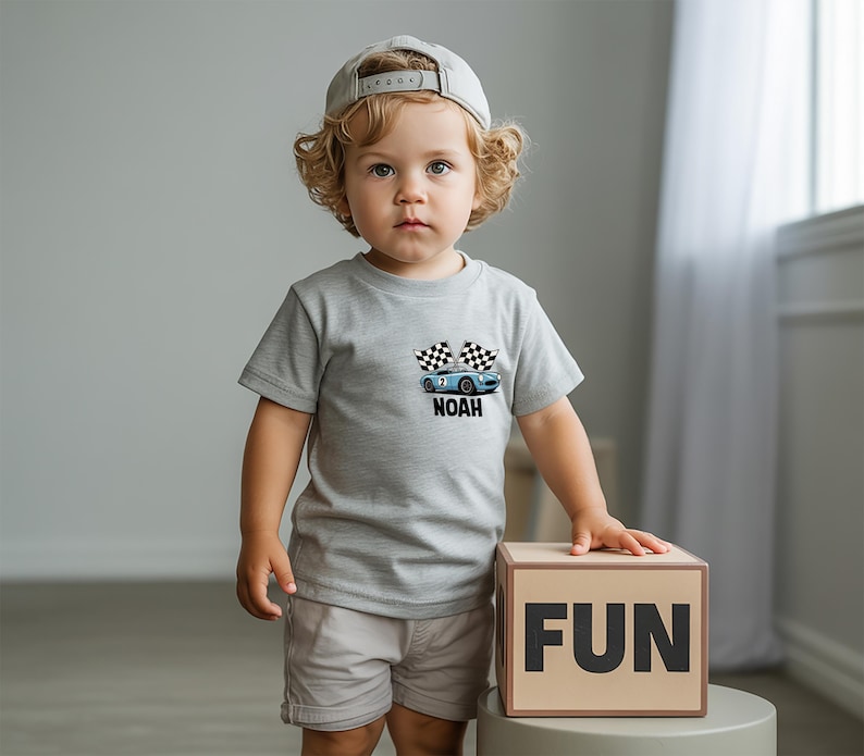 A young child with curly hair wearing a gray t-shirt with a graphic of a car and the word "NOAH" stands next to a cardboard box with the word "FUN" on it.