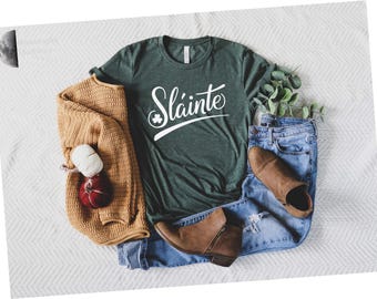 May include: A dark green short-sleeve t-shirt with the white text Sláinte and a small shamrock icon, paired with a mustard yellow knit cardigan, light blue distressed denim jeans, and brown ankle boots, arranged with a small red decorative item.