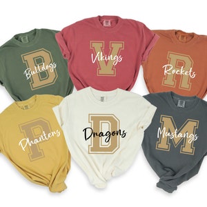 May include: Six different color t-shirts with a large letter and team name printed on the front. The shirts are in the colors green, red, orange, yellow, white, and gray. The letters are all gold.