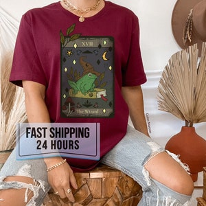 Puede incluir: Una camiseta de color granate con un gr&aacute;fico de una carta de tarot que presenta una rana con un sombrero de mago. La carta est&aacute; etiquetada como "The Wizard" y tiene el n&uacute;mero "XVII" en la esquina superior derecha. La camiseta tiene una etiqueta que dice "FAST SHIPPING 24 HOURS".