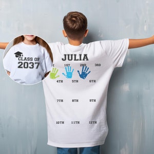 K&ouml;nnte beinhalten: Wei&szlig;es T-Shirt mit dem Text "CLASS OF 2037" auf der Vorderseite und dem Text "JULIA" sowie Handabdr&uuml;cken, die jede Klasse von der 1. bis zur 12. Klasse repr&auml;sentieren, auf der R&uuml;ckseite.
