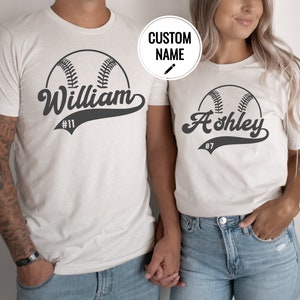 May include: Two white t-shirts with a baseball graphic and personalized names. The shirt on the left says "William #11" and the shirt on the right says "Ashley #7".